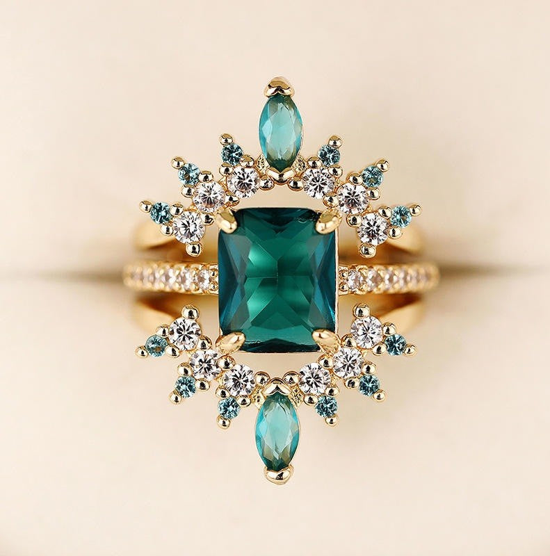 A gold ring featuring a large rectangular emerald-cut green center stone, surrounded by marquise-cut and round clear accent stones in blue-green and white, set against a soft beige background.
