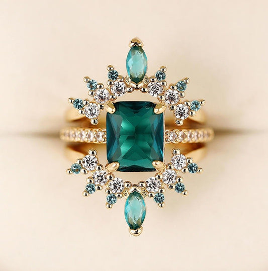 A gold ring featuring a large rectangular emerald-cut green center stone, surrounded by marquise-cut and round clear accent stones in blue-green and white, set against a soft beige background.