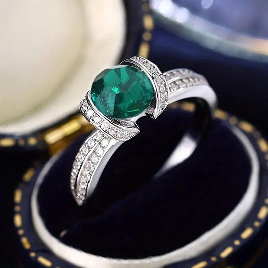 A silver ring featuring a large round green emerald-cut gemstone, accented with small clear stones along the band, displayed in a dark velvet ring box.