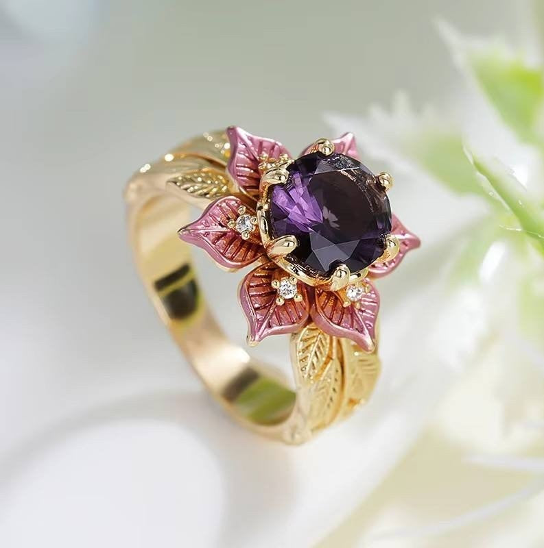 A gold ring designed as a flower, featuring a large dark purple central gemstone surrounded by pink and gold enamel petals with small clear accent stones, set against a soft white background.