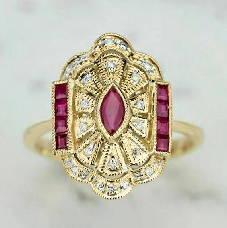 A vintage-style gold ring featuring a central marquise-cut pink gemstone, flanked by rectangular pink stones and surrounded by small clear stones in an ornate filigree setting.