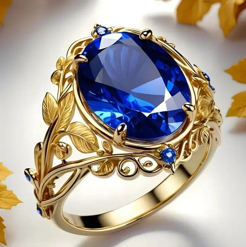 An ornate gold ring featuring a large oval-cut deep blue gemstone in a leafy filigree setting, with smaller blue accent stones, photographed on a light background with scattered yellow leaves.