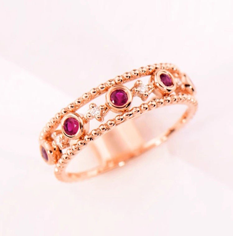A rose gold double-band ring featuring round-cut deep pinkish-red gemstones and small clear accent stones, set against a clean white background.