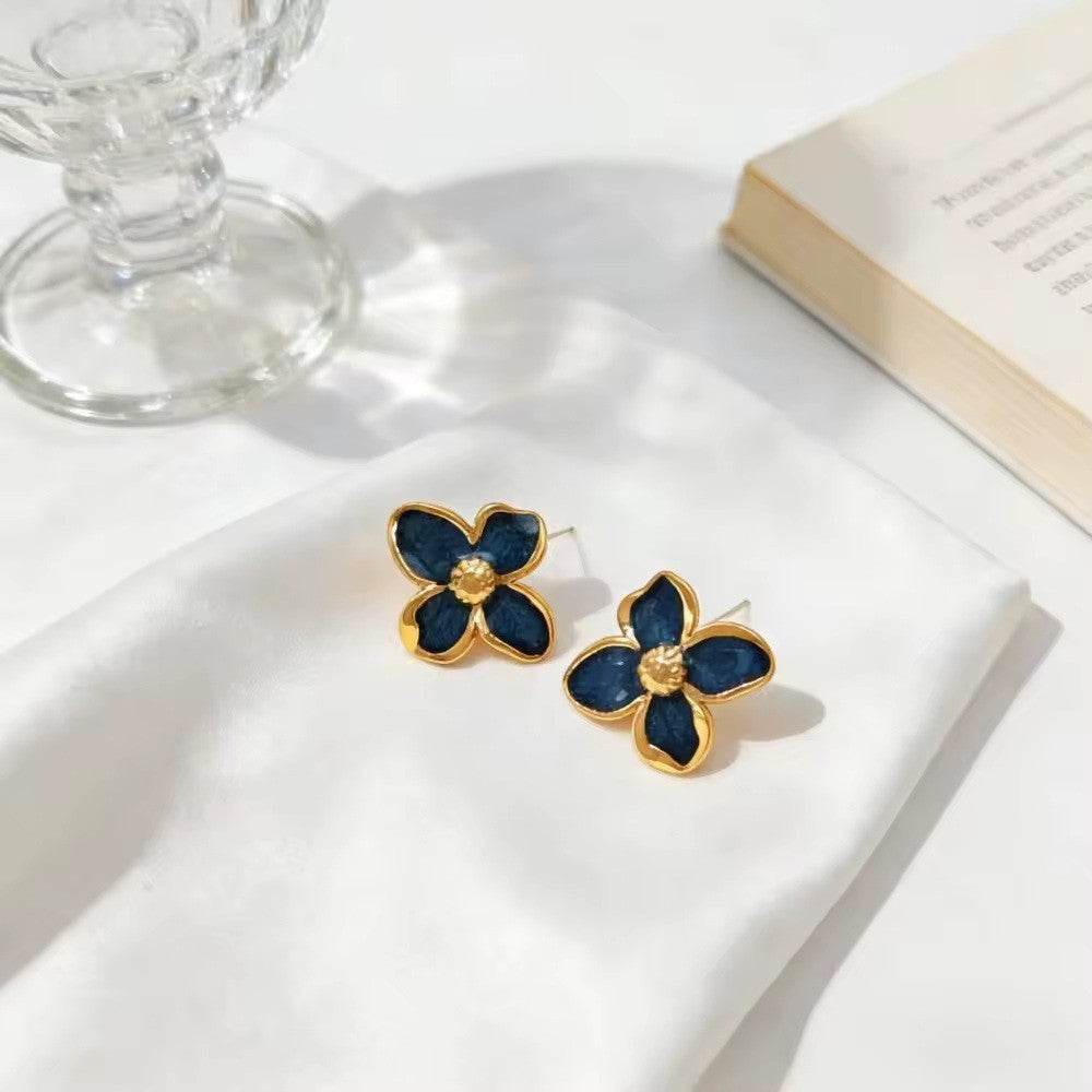 A pair of gold-tone flower-shaped earrings with dark blue enamel petals and a small clear stone in the center, displayed on a white satin cloth with a glass candle holder and an open book in the background.