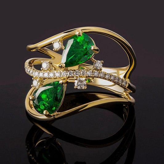 A gold ring featuring two large emerald-cut green gemstones and a curved band paved with small clear stones, photographed on a glossy black reflective surface.