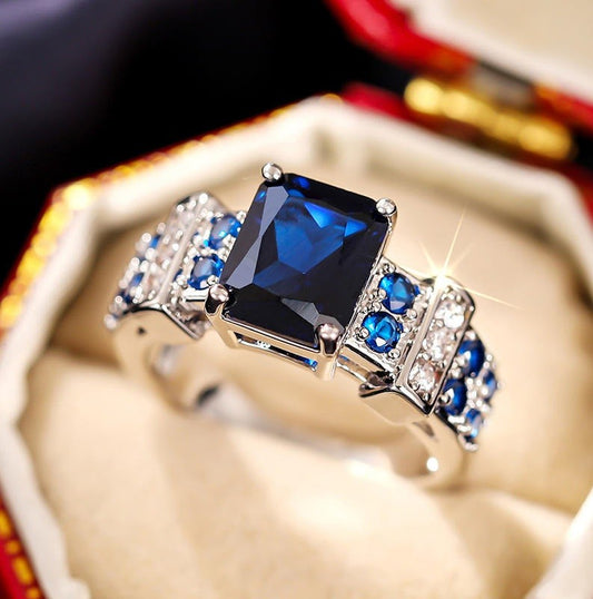 A silver ring featuring a large rectangular dark blue gemstone and smaller blue and clear stones, displayed inside a beige ring box.