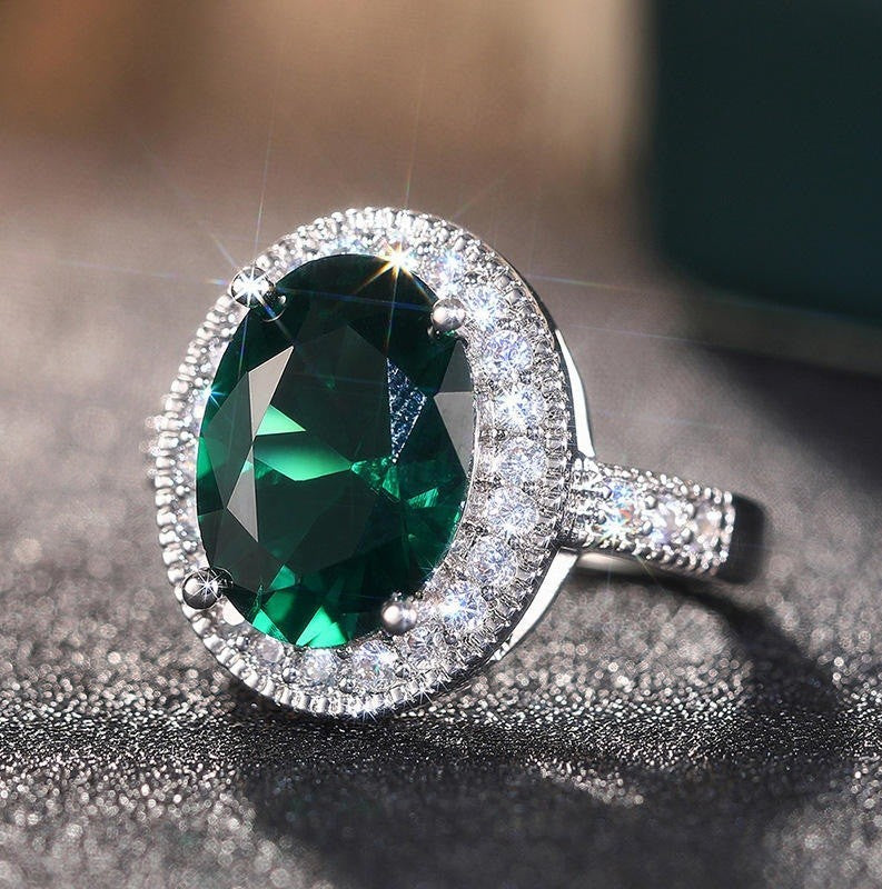 An oval halo ring featuring a large green emerald-cut center stone surrounded by a halo of small clear stones, set on a silver-toned band and photographed close-up on a dark textured surface.