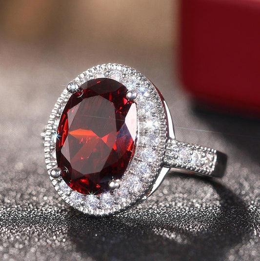 An oval halo ring featuring a large faceted deep red gemstone in a silver-toned setting, surrounded by a halo of small clear stones, resting on a dark textured surface.