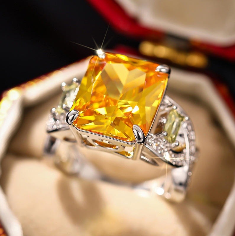 A silver ring featuring a large, sparkling yellow cushion-cut gemstone, displayed inside an open ring box with a blurred background.