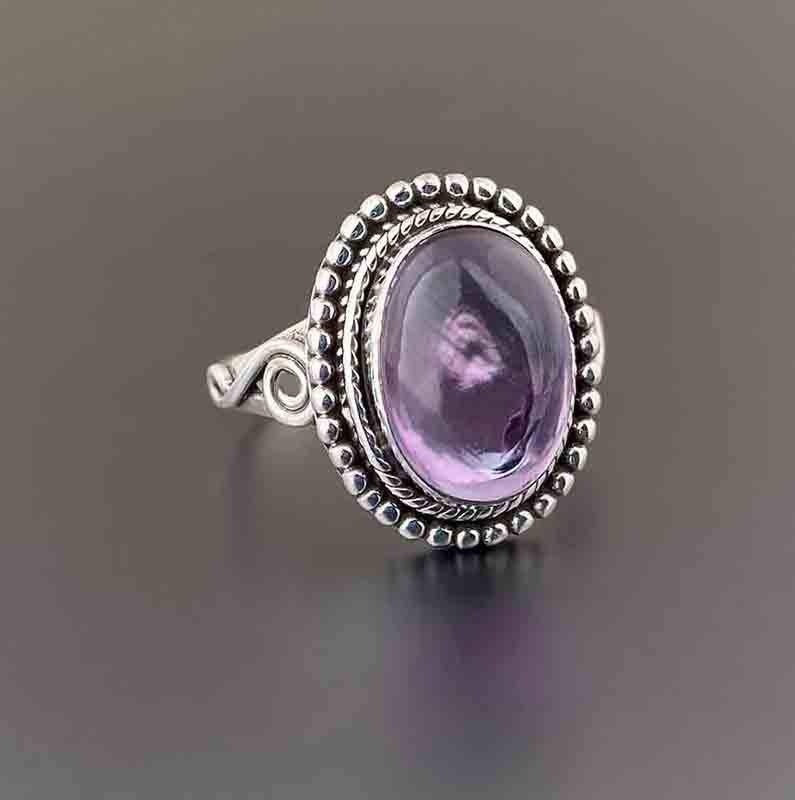 A silver ring featuring an oval cabochon amethyst gemstone, set in a textured bezel with a twisted rope border and a decorative band, shown on a dark gray reflective surface.