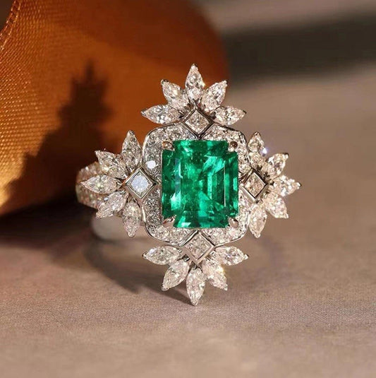 An ornate silver-toned ring featuring a large rectangular green emerald-cut center stone surrounded by multiple round and marquise-cut clear stones in leaf and starburst motifs, photographed on a soft beige surface with a warm brown ribbon in the background.