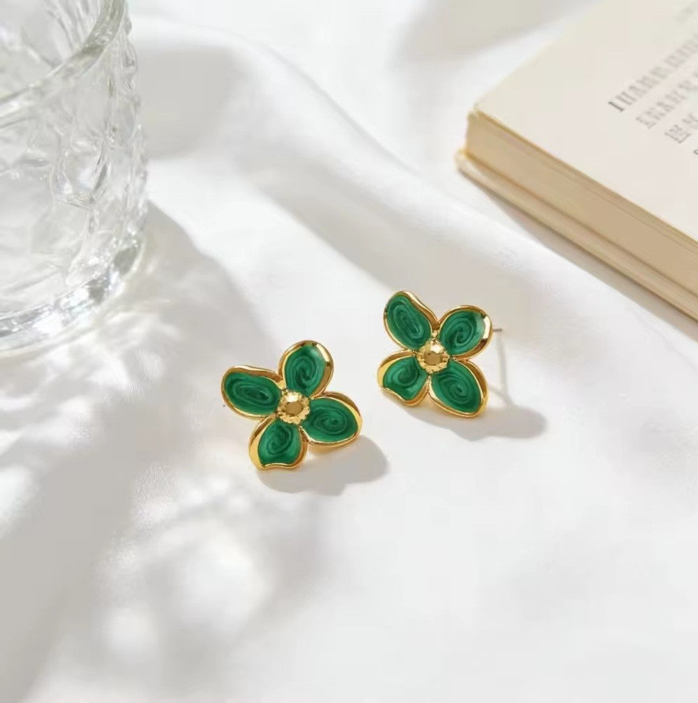 A pair of gold-tone flower stud earrings with green enamel petals, displayed on a white fabric surface next to a glass and a book.