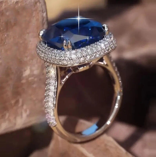 A close-up shot of a silver ring featuring a large oval-cut deep blue gemstone in a prong setting, surrounded by a halo of small pavé-set clear stones and a split shank with matching pavé detailing.