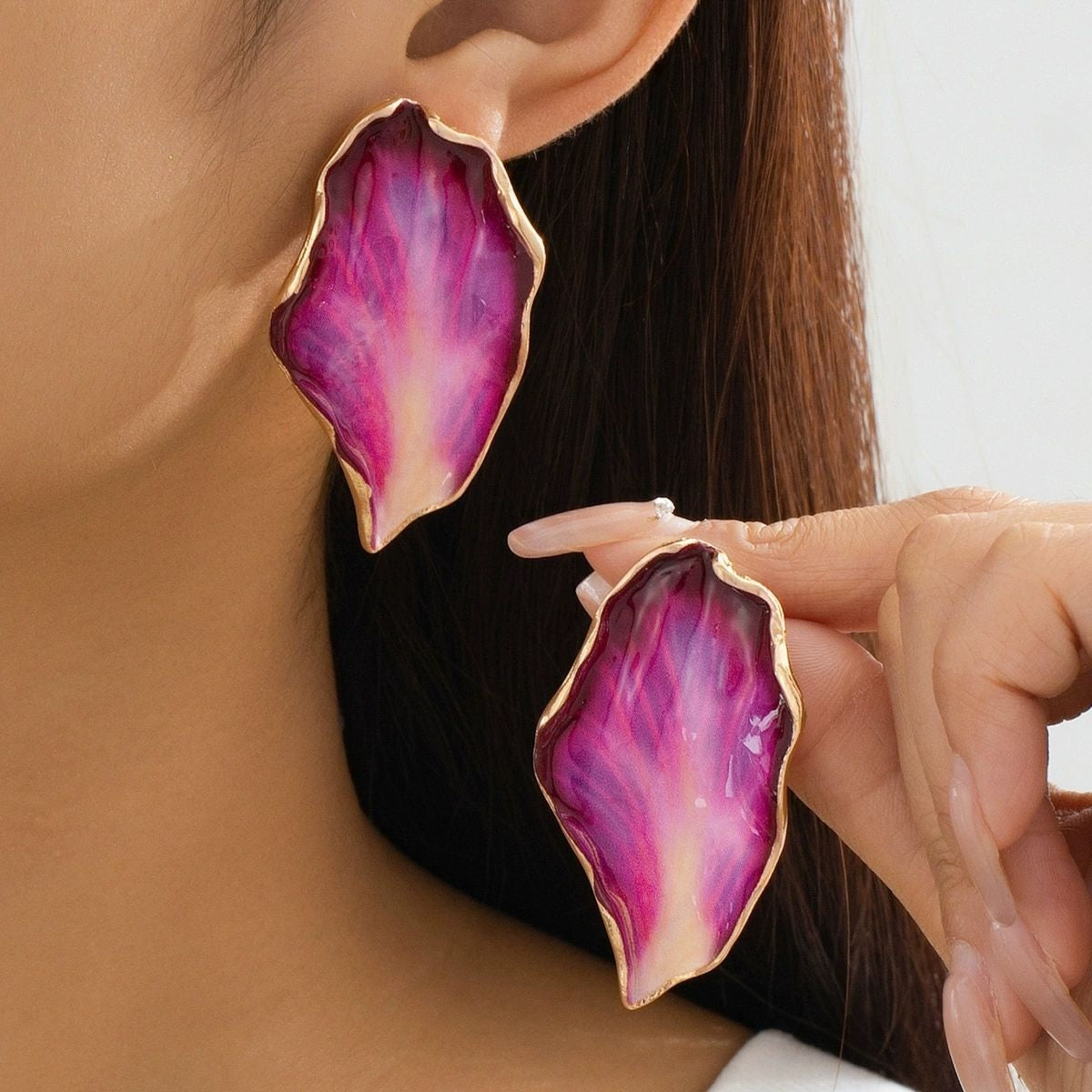 A close-up lifestyle shot of a model wearing and holding a pair of large, organic-shaped drop earrings. The earrings feature a vibrant magenta and purple marbled pattern with a polished gold-tone border.