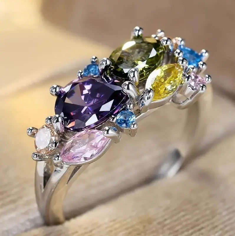 A silver-colored ring featuring a cluster of multicolored gemstones, including a large purple oval, a yellow marquise-cut stone, a pink marquise-cut stone, and smaller blue and green stones, set against a warm beige background.