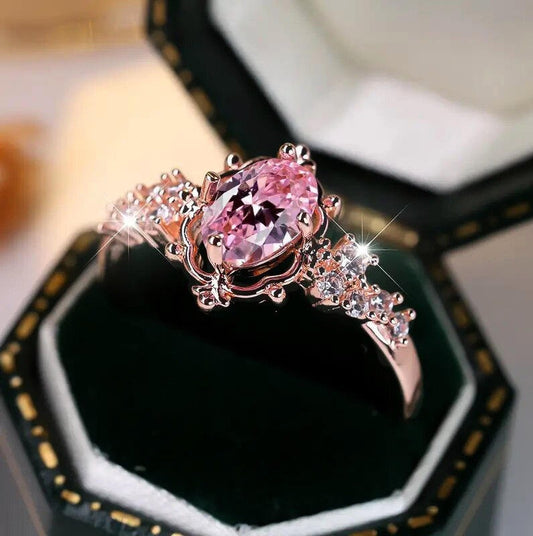 A pink gemstone ring with rose gold metal detailing, displayed in an open black jewelry box.