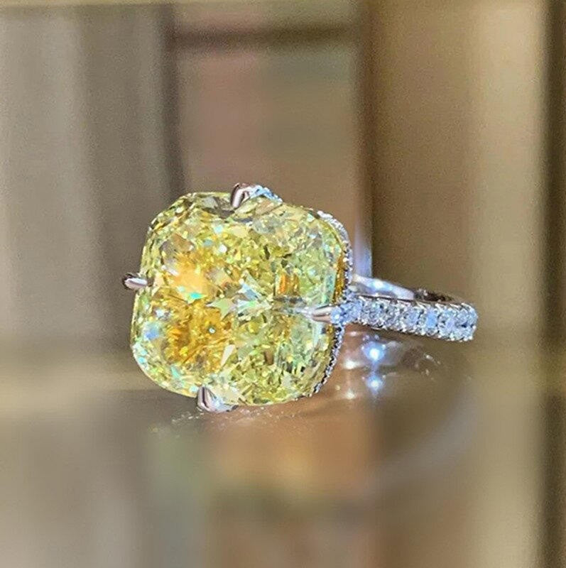 A cushion-cut yellow diamond ring with a silver-toned band set with small pavé diamonds, shown on a reflective surface with a blurred warm-toned background.
