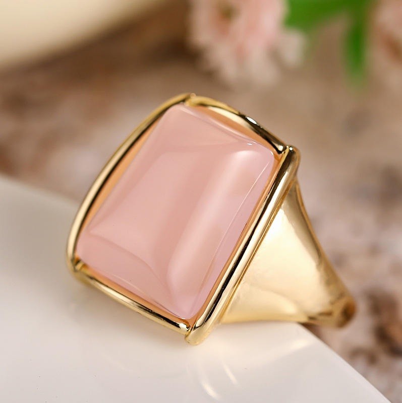 A gold signet ring featuring a large, smooth, rectangular pale pink stone, resting on a white surface with a softly blurred background.
