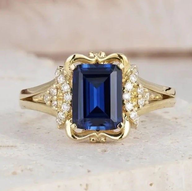 A yellow gold ring featuring a rectangular emerald-cut blue sapphire center stone, accented by small clear stones on the shoulders, set against a soft beige background.