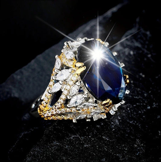A gold and silver ring featuring a large oval-cut dark blue gemstone, surrounded by marquise-cut clear stones and gold accents, set against a dark textured background.