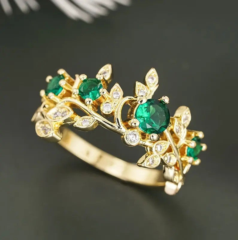 A gold ring designed as a leafy vine, featuring round green emerald-cut gemstones and small clear accent stones, photographed on a dark gray background.