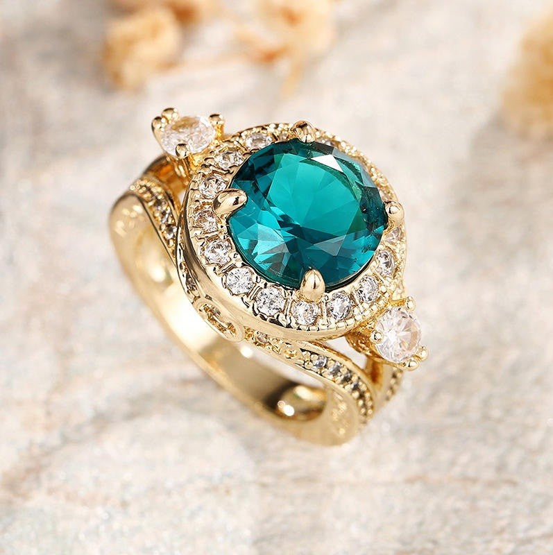 A gold ring featuring a large oval-cut teal blue gemstone surrounded by a halo of small clear stones, with additional clear accent stones on the band, photographed on a light textured surface.