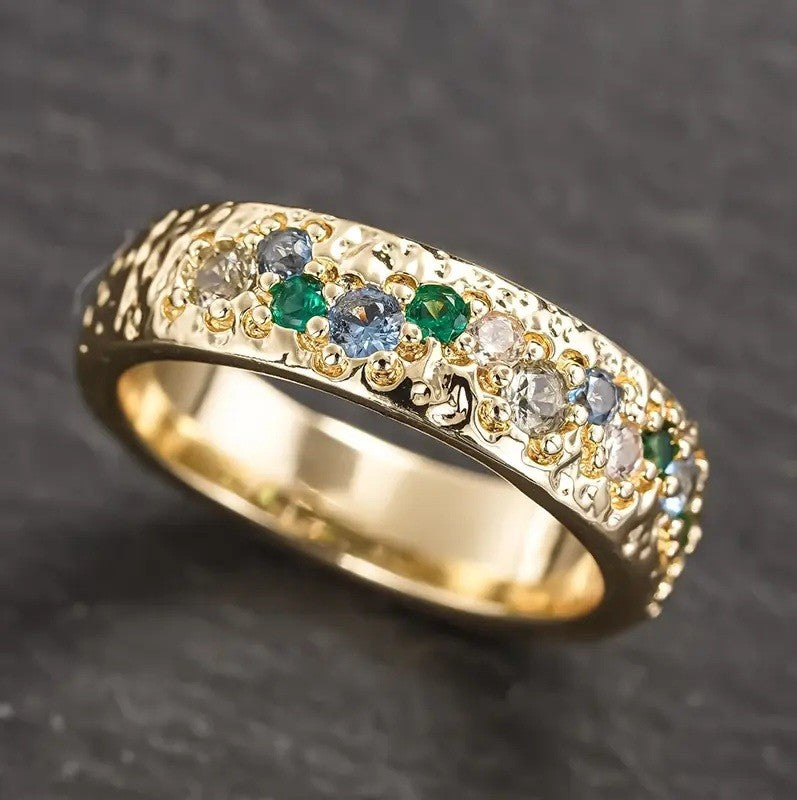A gold eternity ring featuring a row of round-cut clear, light blue, and green gemstones on a textured band, set against a dark gray slate background.