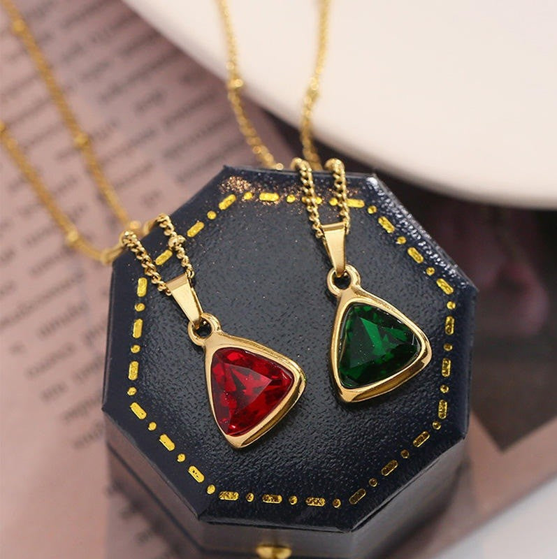 Two gold-tone triangle pendant necklaces with faceted red and green gemstones, displayed on a black hexagonal jewelry box.