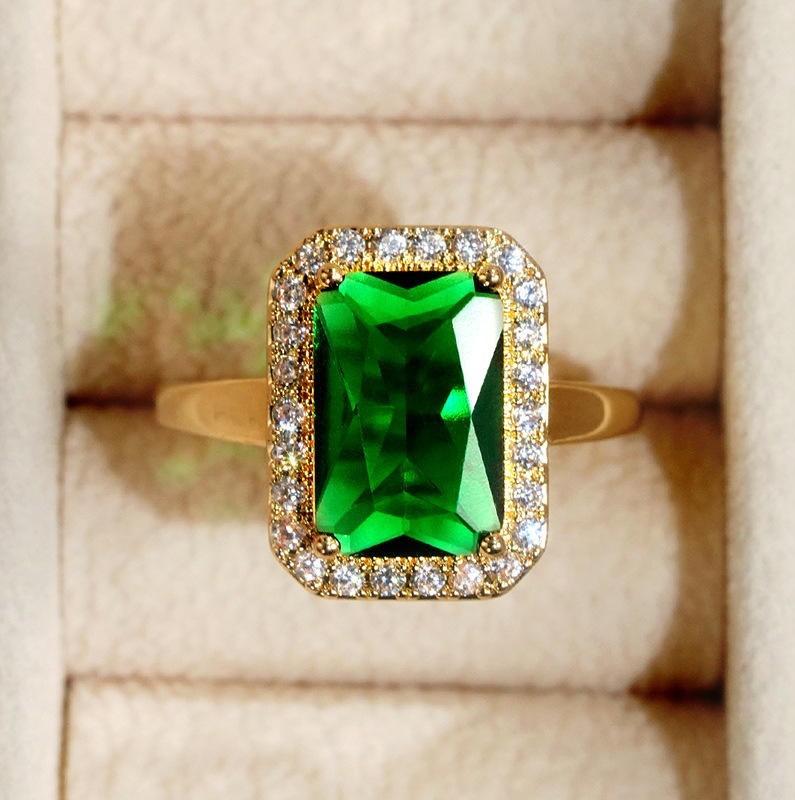 A yellow gold ring featuring a rectangular emerald-cut green gemstone surrounded by a double halo of small clear stones, displayed on a beige ring box.