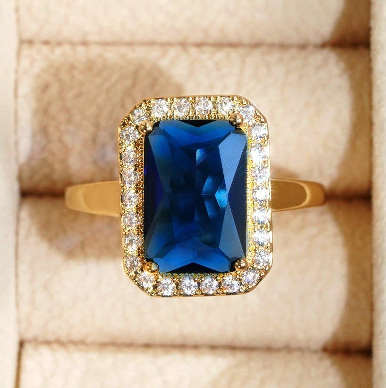 A yellow gold ring featuring a rectangular blue gemstone surrounded by a halo of small clear stones, displayed on a beige ring box.