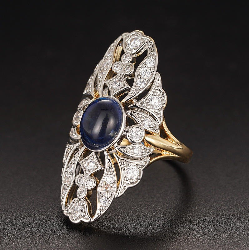 An ornate Art Deco style ring featuring a central oval blue sapphire surrounded by pavé-set diamonds in silver-toned metal, with a yellow gold band and matching gold accents, set against a dark textured background.