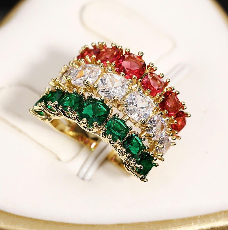 A gold-plated triple-row ring featuring green, white, and red gemstone rows, displayed on a white ring stand.