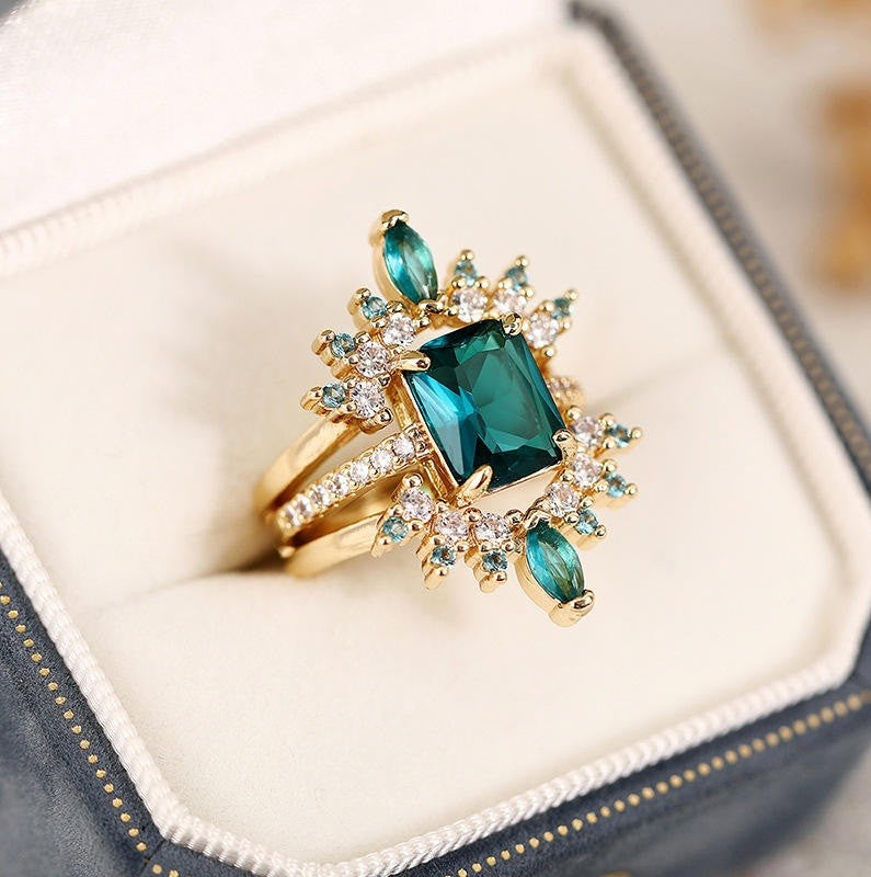 A gold ring with a large emerald-cut green center stone and smaller green marquise stones, accented with clear crystals, displayed in a cream-colored ring box.