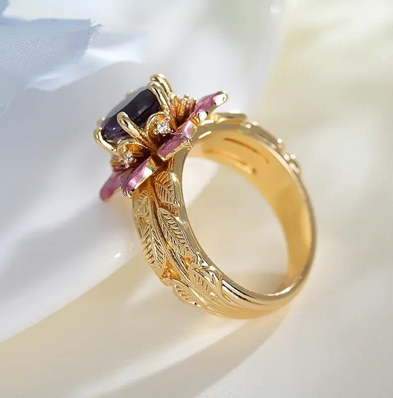 A gold ring featuring a flower-shaped setting with a dark purple rectangular gemstone and pink gemstone accents, resting on a soft white fabric background.