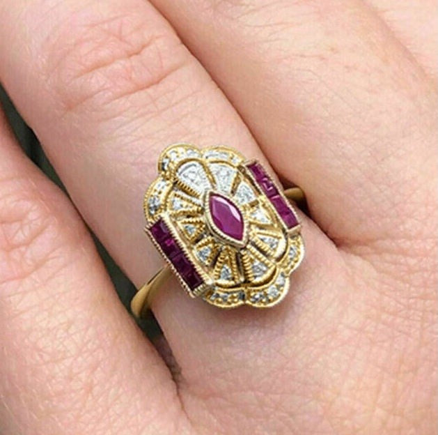 A close-up of a vintage-style Art Deco ring with a yellow gold setting, featuring a central marquise-cut ruby and side rectangular rubies, accented with small clear stones, worn on a finger.