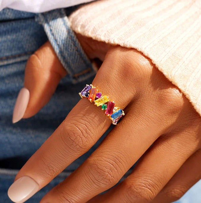 A close-up of a hand wearing a silver ring set with colorful rectangular gemstones in a rainbow sequence, styled with a cream ribbed sweater and blue jeans.