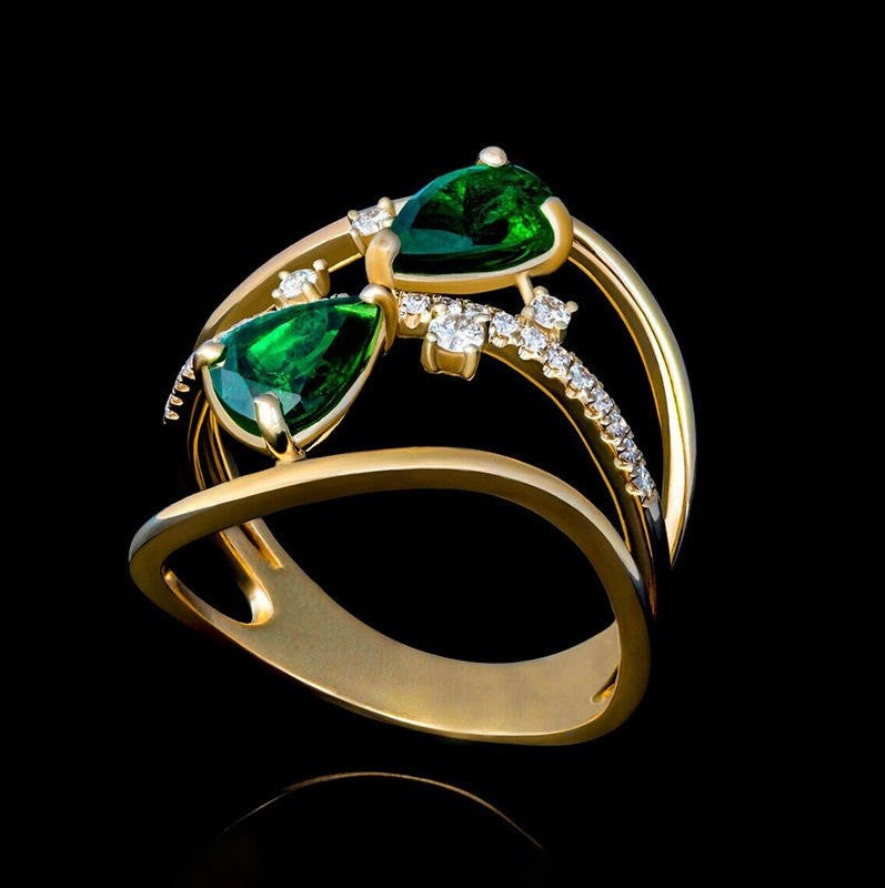 A gold ring featuring two large green teardrop-shaped gemstones and a curved band accented with small clear stones, shown on a black background.