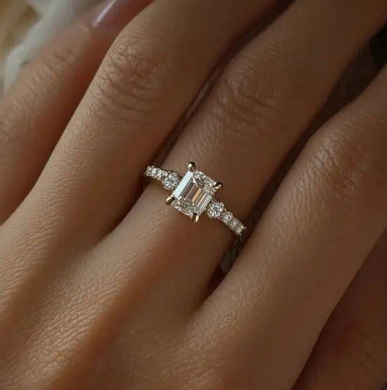 A close-up of a silver engagement ring with an emerald-cut center stone and smaller side stones, worn on a finger.
