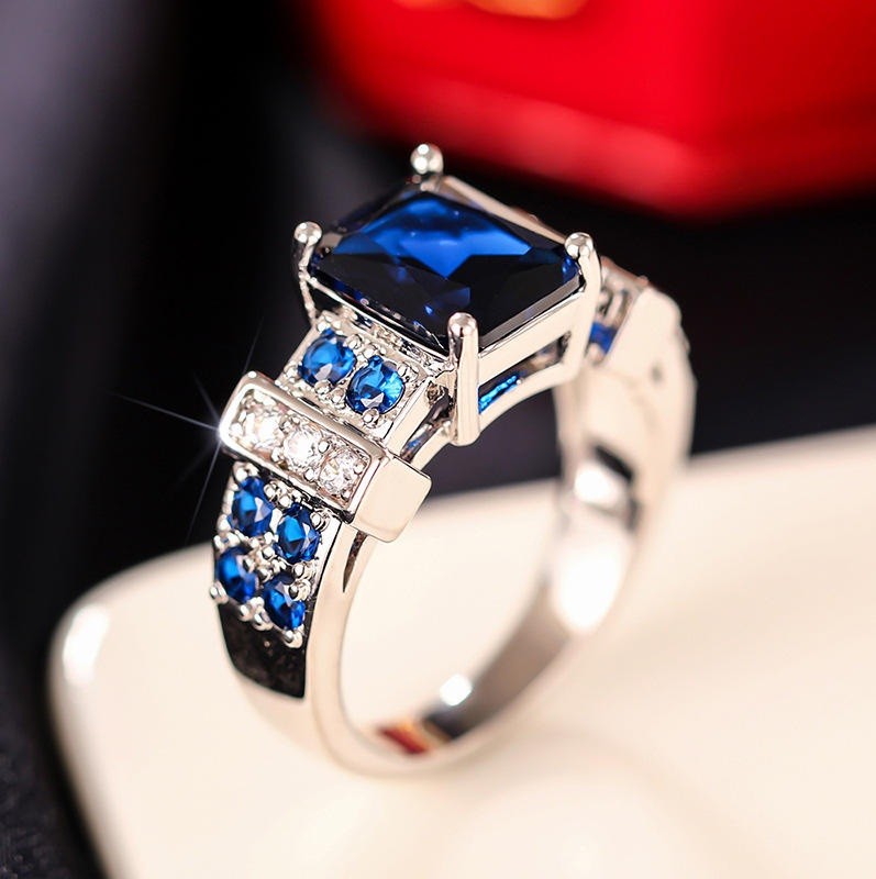 A silver-toned ring featuring a large rectangular deep blue gemstone, accented with smaller round blue and clear stones on the shoulders, displayed on a cream-colored surface with a blurred red and black background.