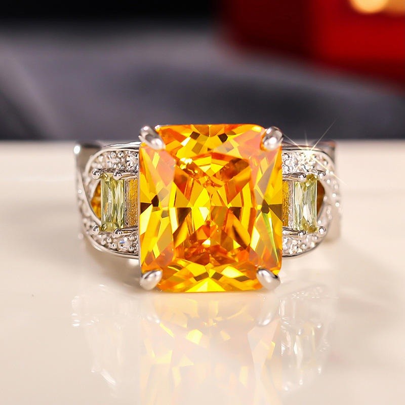 A silver-toned ring featuring a large, vibrant yellow-orange cushion-cut center stone, flanked by two small light green baguette-cut stones and a halo of tiny clear accent stones on a white reflective surface.