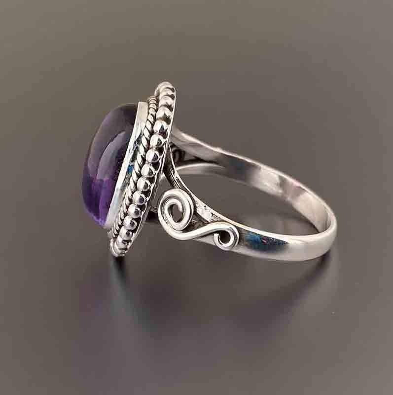A sterling silver ring featuring a bezel-set oval purple amethyst gemstone, shown on a dark gray reflective surface.