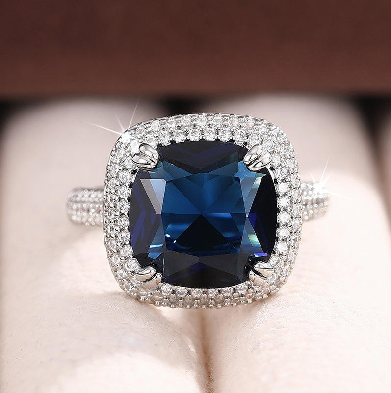 A cushion-cut blue sapphire ring with a diamond halo in a silver-toned setting, displayed on a soft cream-colored ring box.
