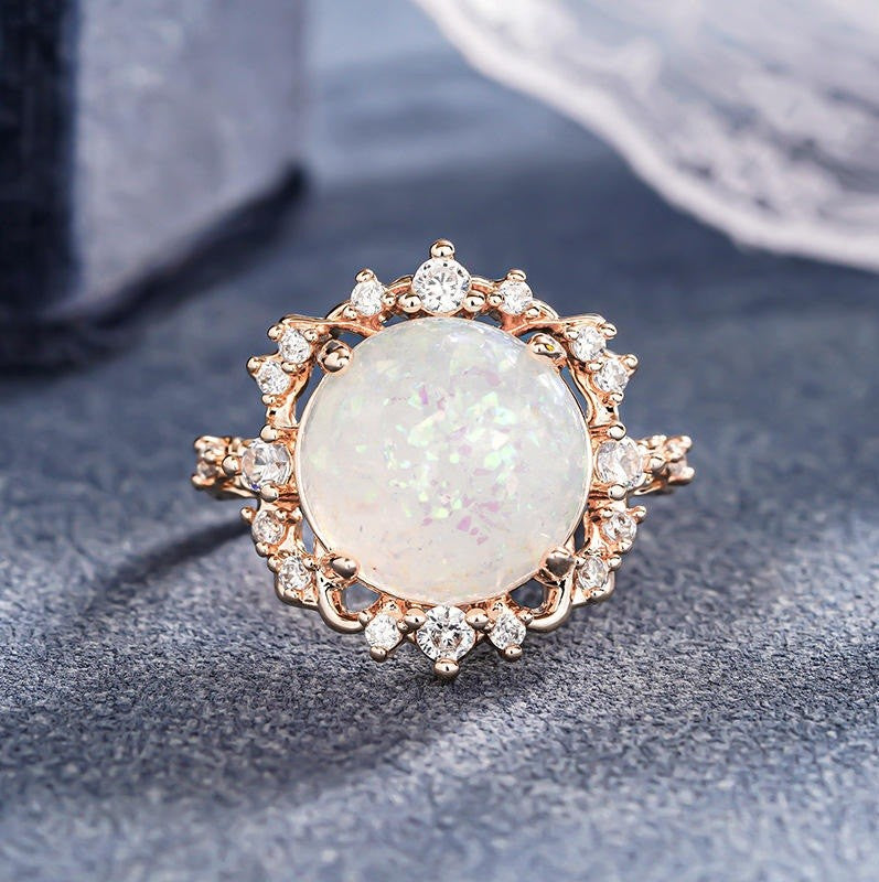 A rose gold ring featuring a large oval white opal center stone surrounded by small clear accent stones, displayed on a textured dark gray surface.