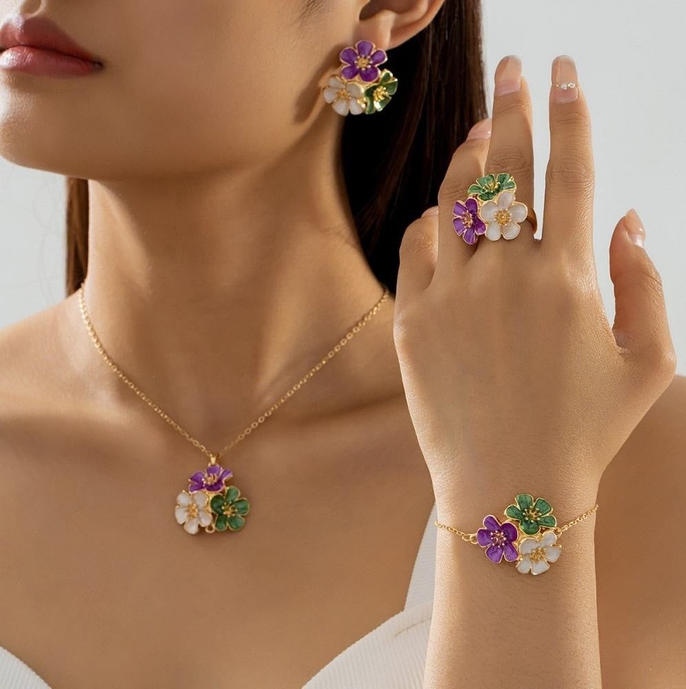 A close-up lifestyle shot of a model wearing a matching gold jewelry set with colorful floral pendants, including a necklace, ring, and bracelet. The jewelry features purple, white, and green enamel flowers on delicate gold settings.