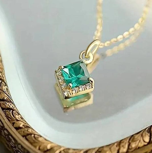 A gold pendant necklace featuring a square-cut green gemstone surrounded by small clear stones, resting on a reflective white surface with an ornate gold frame visible at the edge.