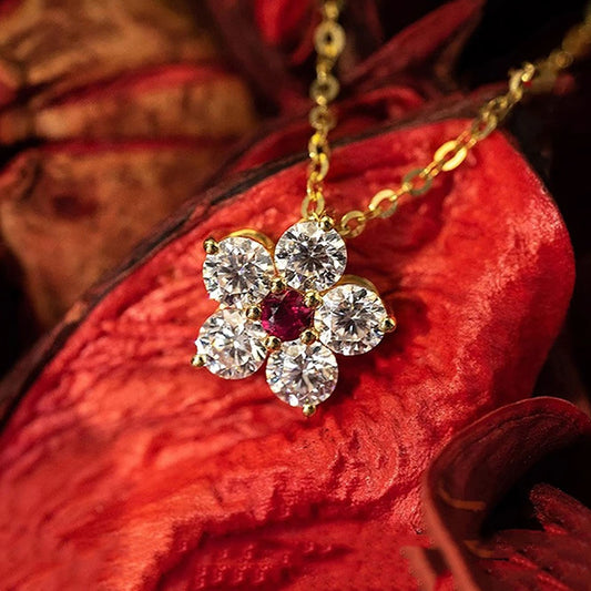 A gold flower-shaped pendant necklace with five clear round-cut stones and a small red center stone, displayed on a textured red flower petal.