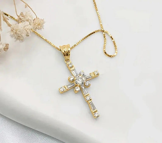 A gold cross pendant necklace adorned with clear baguette and round crystals, resting on a white fabric surface with dried flowers in the corner.