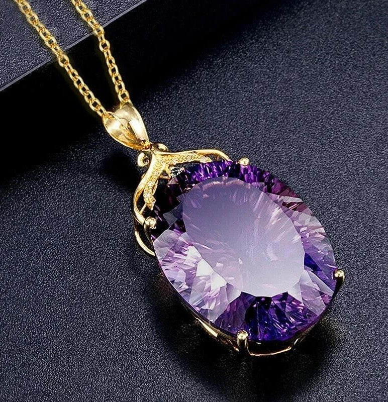 A gold pendant necklace featuring a large, faceted oval purple gemstone, displayed on a textured black background.