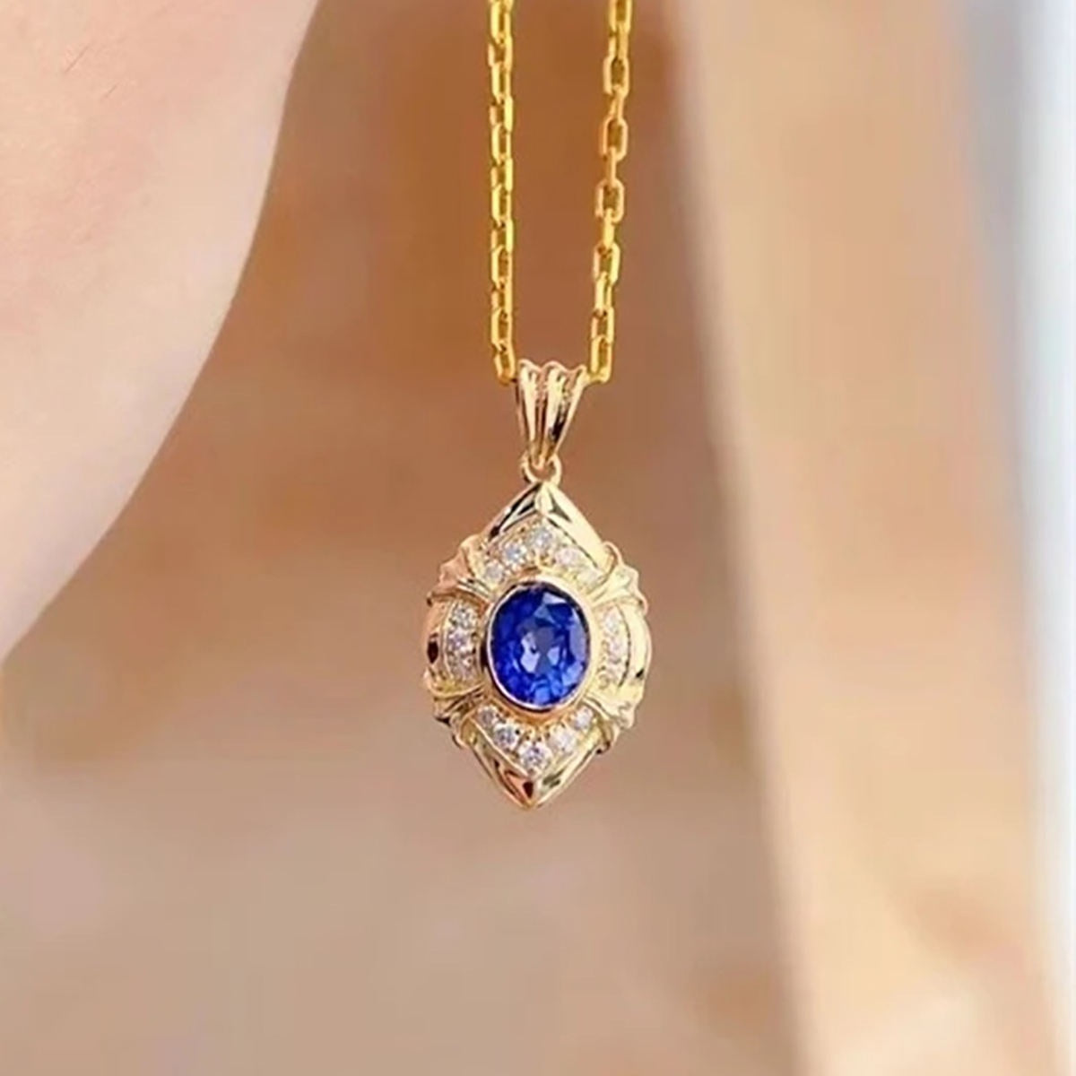 A gold pendant necklace with a central round blue gemstone and small clear accent stones, shown hanging against a softly blurred skin-toned background.