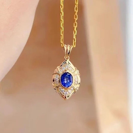 A gold pendant necklace with a central round blue gemstone and small clear accent stones, shown hanging against a softly blurred skin-toned background.